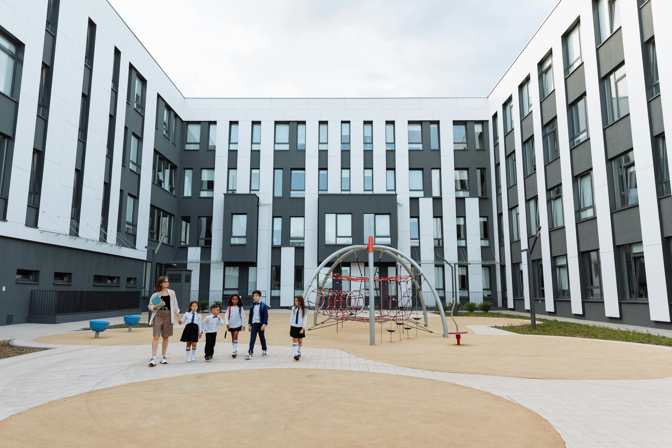 Group of students and teacher walking in a school courtyard with modern architecture and playground.